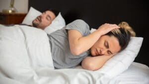 Woman covering her ears in bed while her partner sleeps with his mouth open snoring