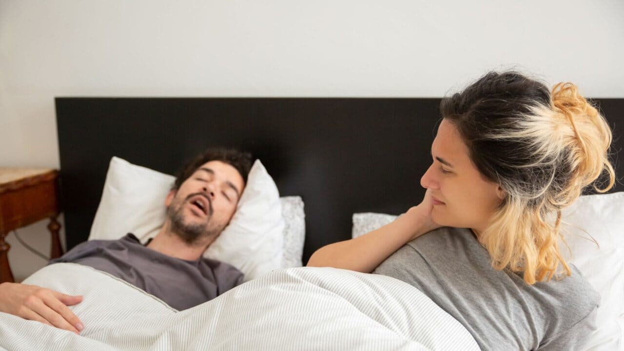 Man snoring with mouth open in bed while his partner lies awake beside him, looking at him