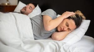 Partner snoring in bed while a woman covers her ears with her hands
