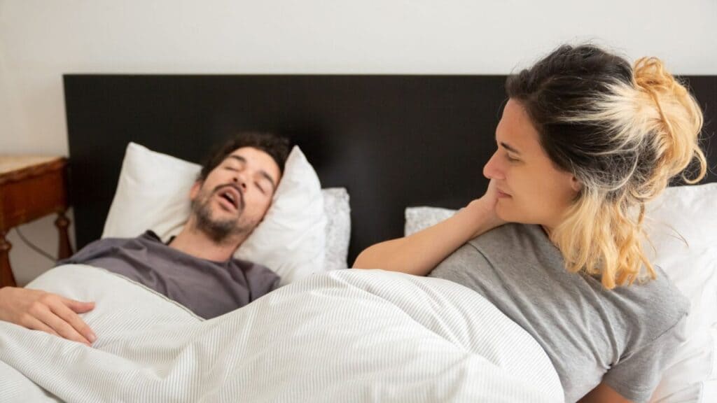 Man snoring on his back while his partner lies awake beside him