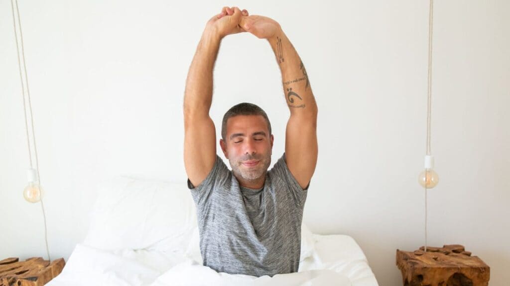 Man stretching his arms overhead while sitting up in a bright bedroom
